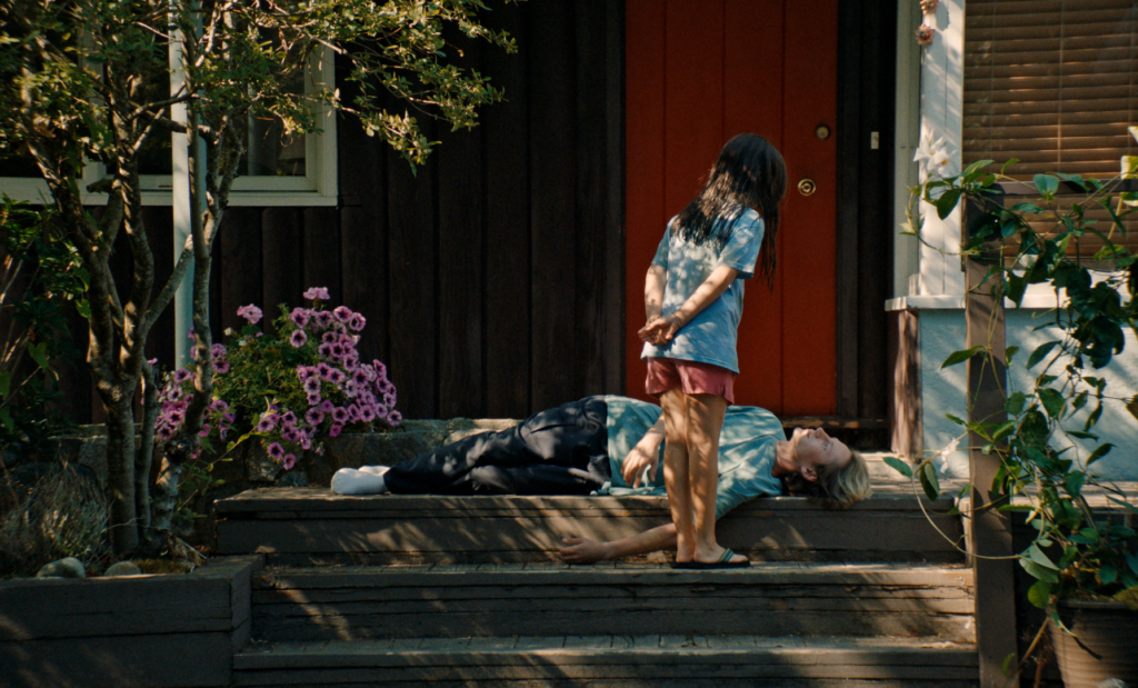 A small girl with dark hair in a blue t-shirt faces away from the camera, looking down at her older brother who is lying on the steps of their home with a red door.